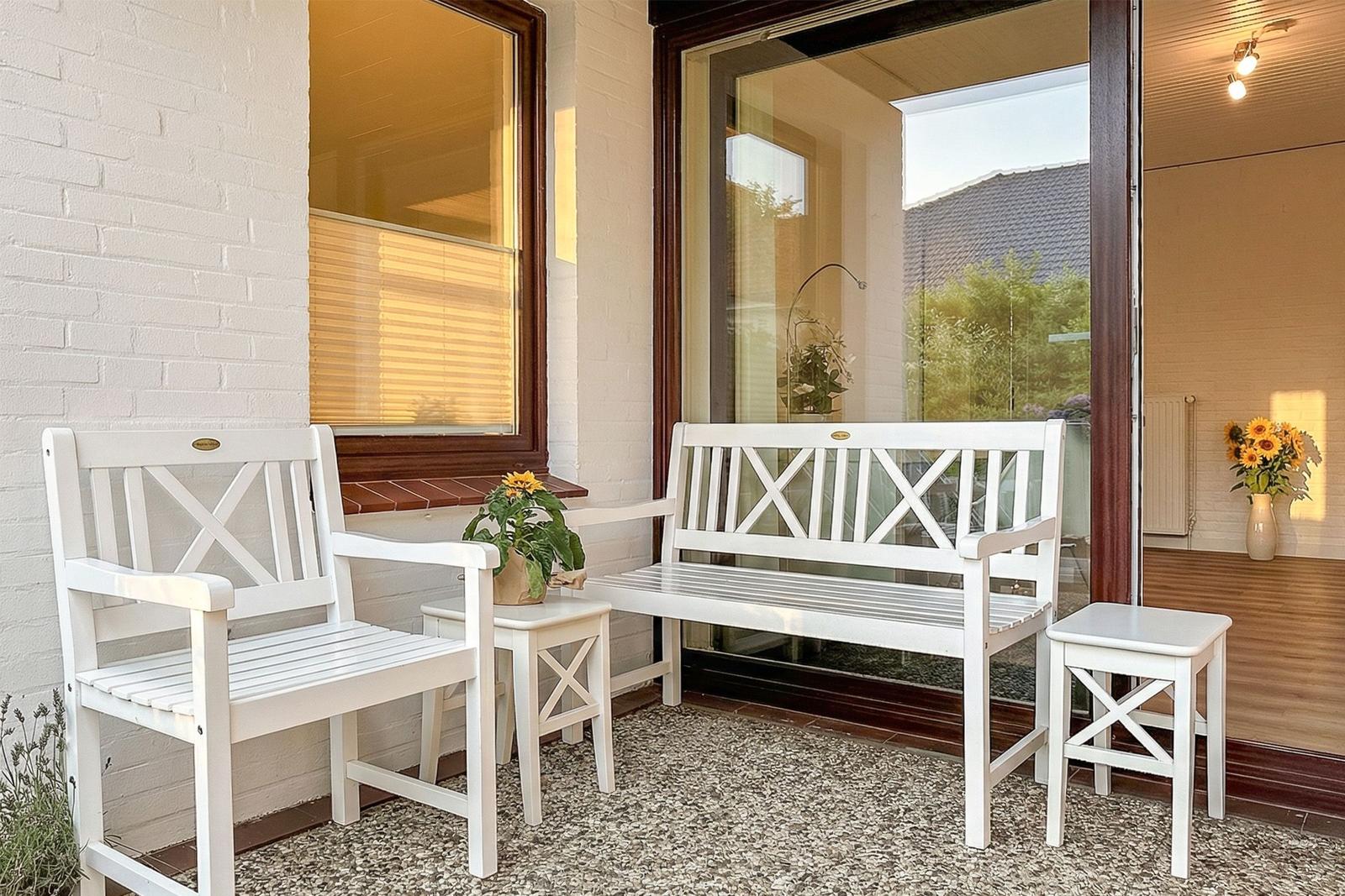 Terrasse mit weißen Sitzgelegenheiten und Blumen vor Glasfront.