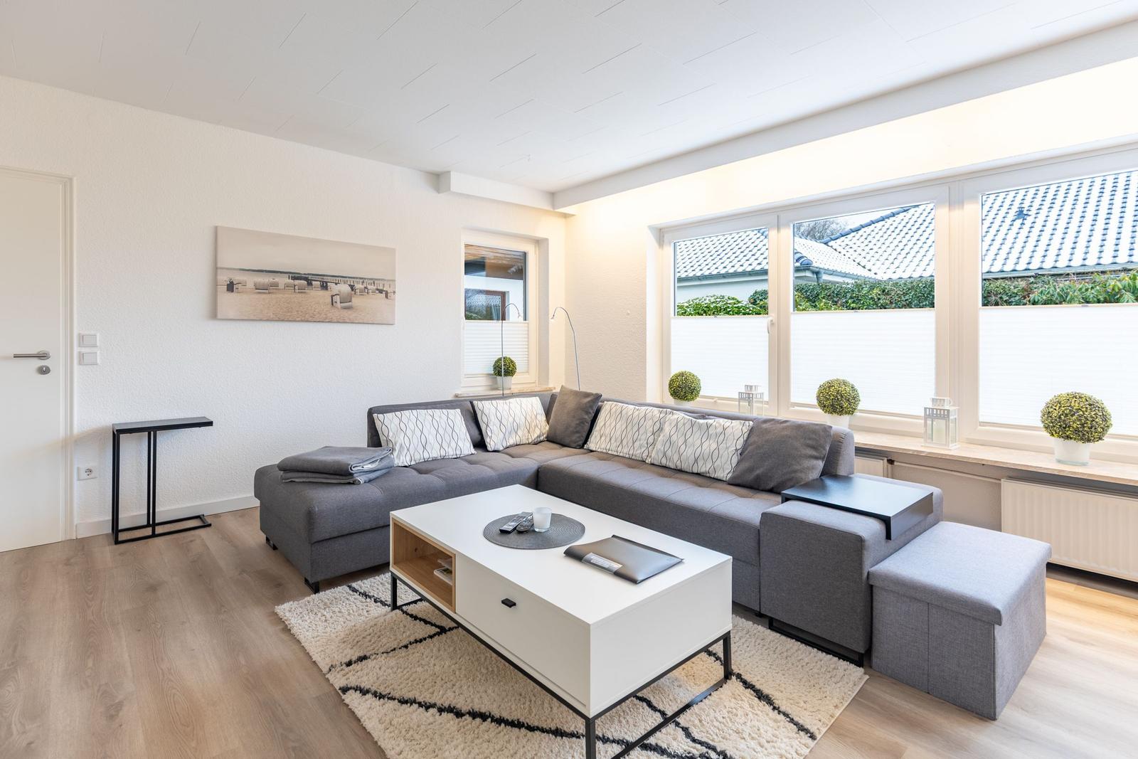 Wohnzimmer mit grauem Sofa, weißen Fenstern und Holzboden.