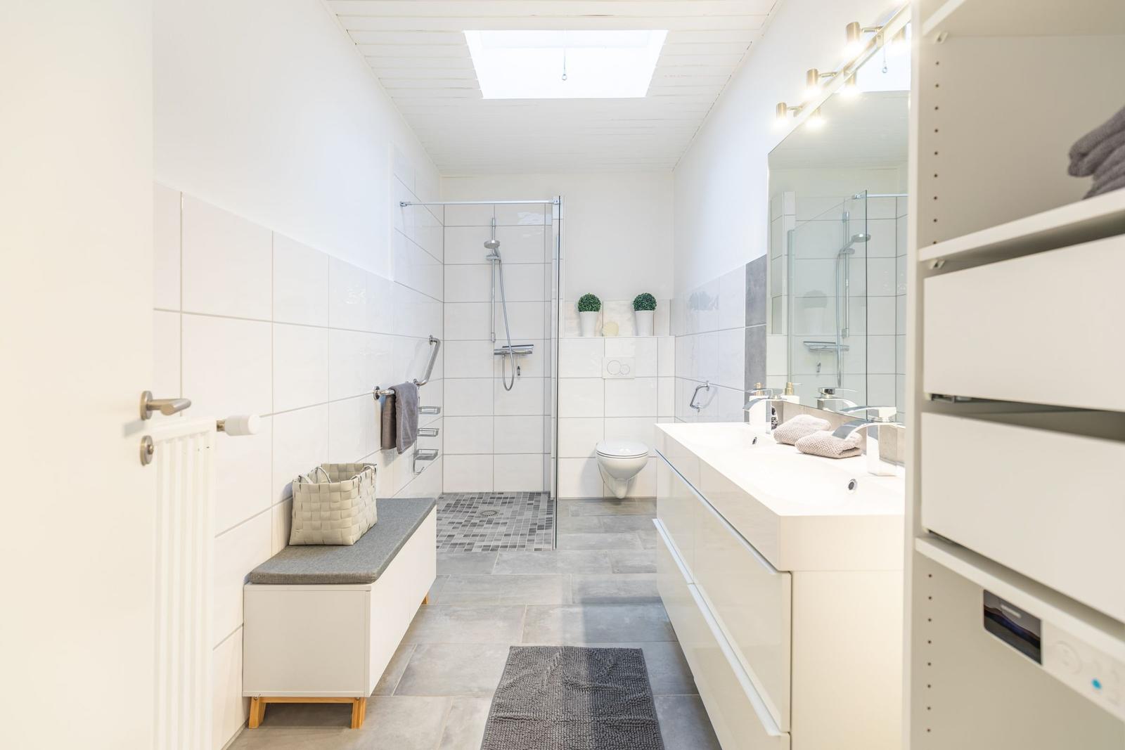 Badezimmer mit Dusche, Waschbecken und Toilette. Weiße Fliesen und graue Bodenfliesen.