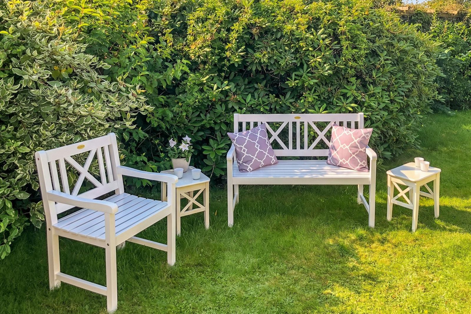 Weißer Gartenbereich mit Bank, Sessel und Tischen. Grüne Hecke im Hintergrund.
