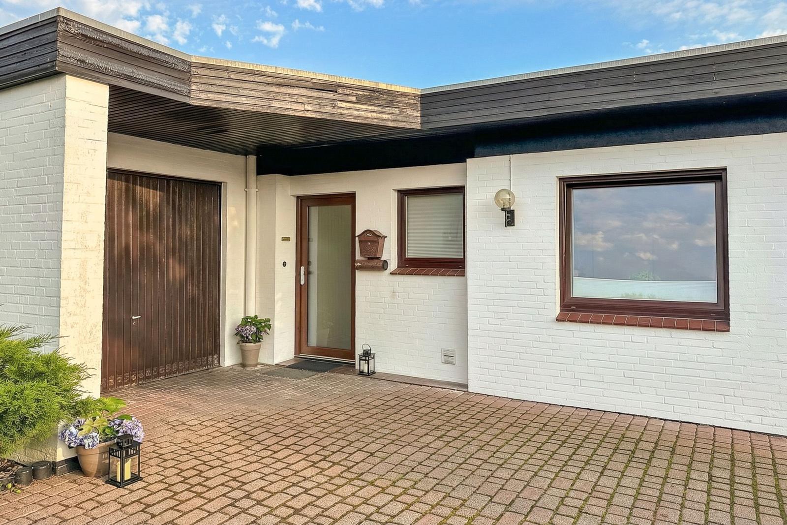 Haus mit weißen Wänden, braunen Fensterrahmen und Garage. Vorgarten mit Pflanzen und Steinplatten.