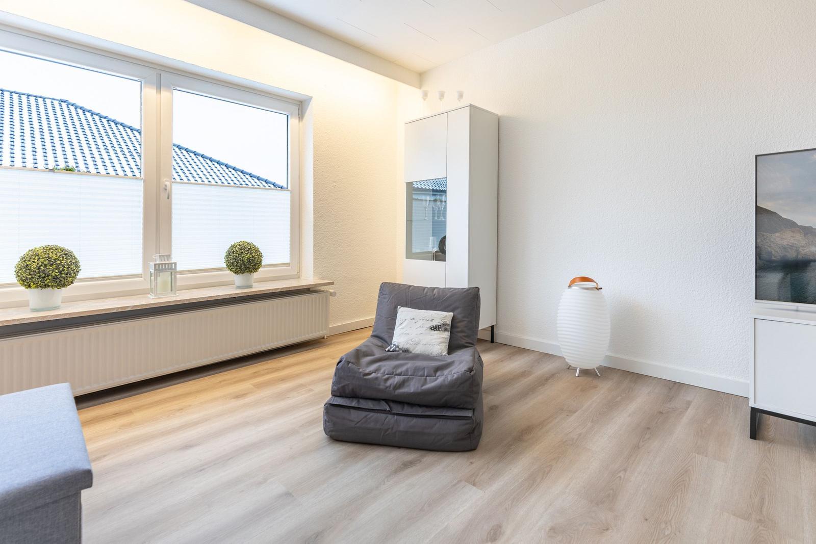 Ein Raum mit grauem Sitzkissen, weißen Wänden und Holzboden. Fenster mit Pflanzen und Schrank.