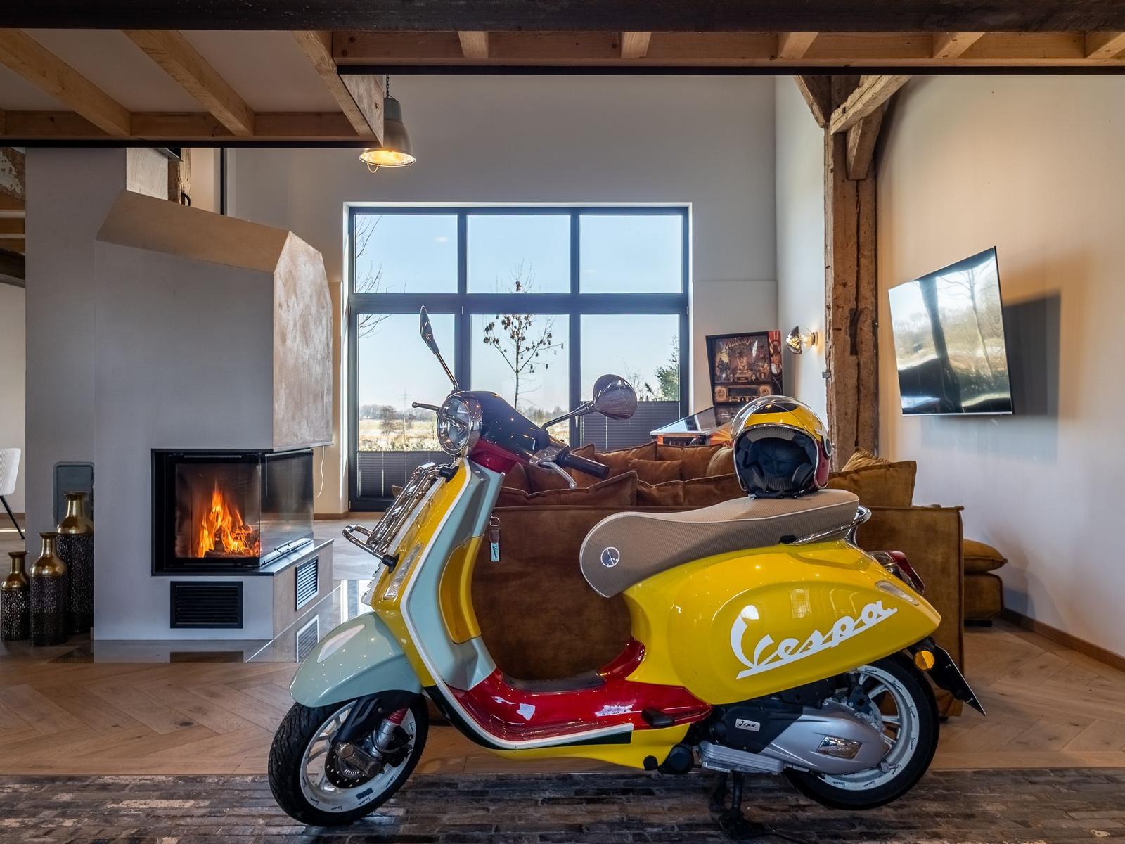 Ein gelbes Vespa-Motorrad steht im Wohnzimmer vor einem Kamin und großen Fenstern.