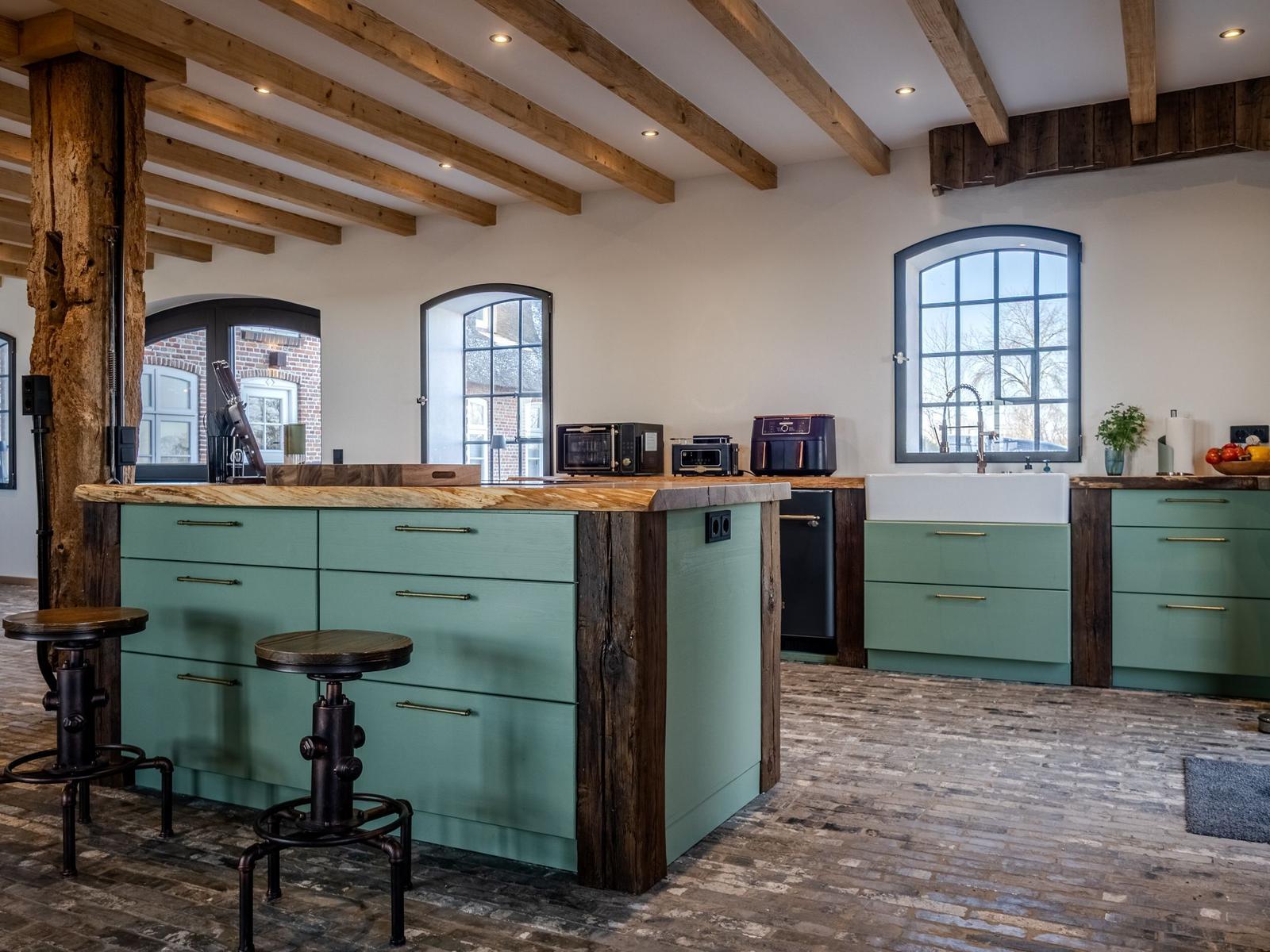 Küche mit grünen Schränken, Holzdecke und Steinboden. Barhocker und Fenster sind sichtbar.