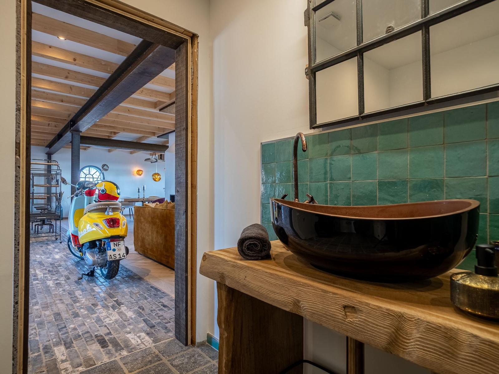 Bad mit Holzwaschtisch, grüner Fliesenwand und Spiegel. Durchgang zeigt Wohnbereich mit Motorrad.