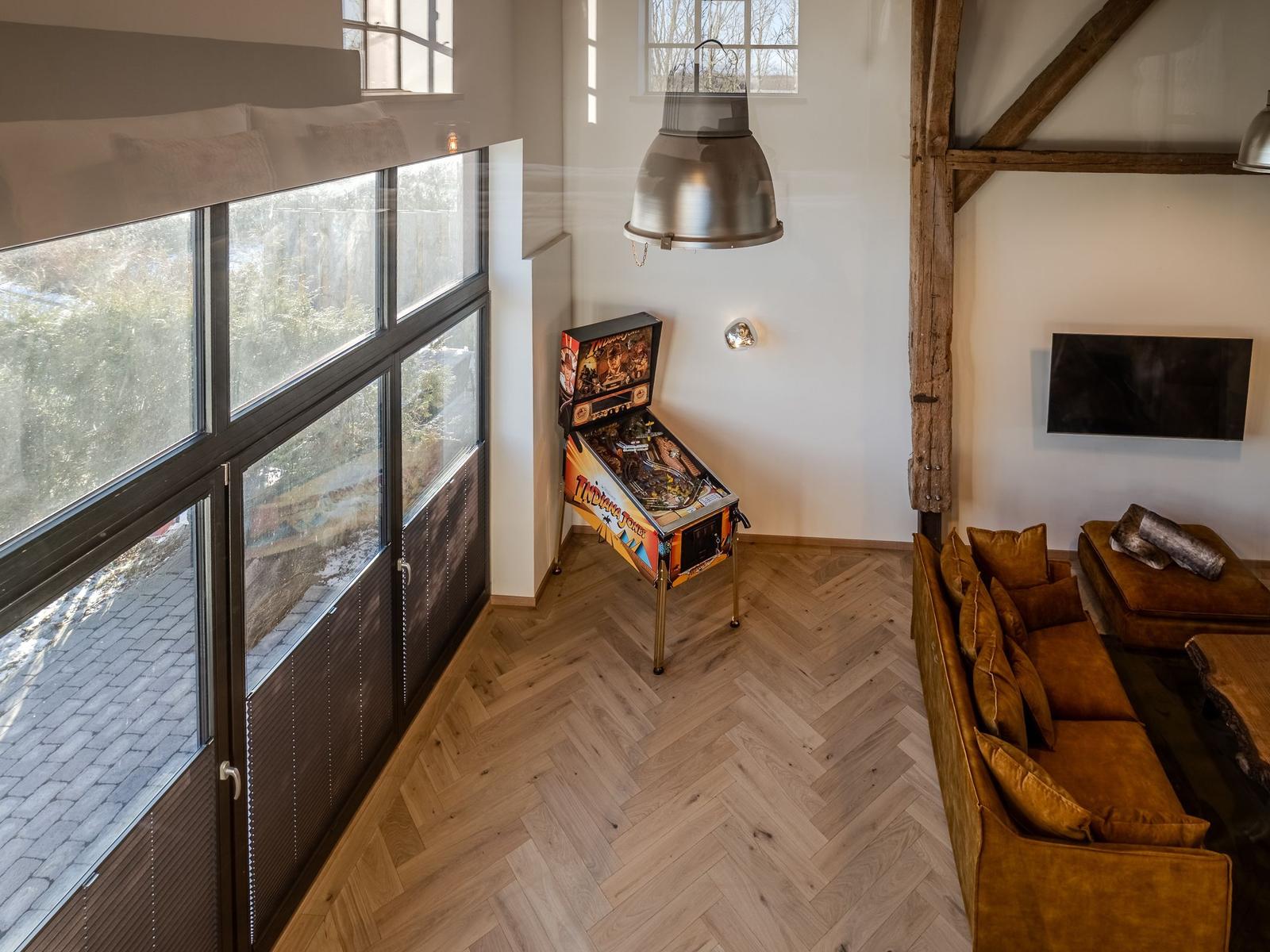 Wohnraum mit Holzboden, Sofa, Pinball und großen Fenstern.