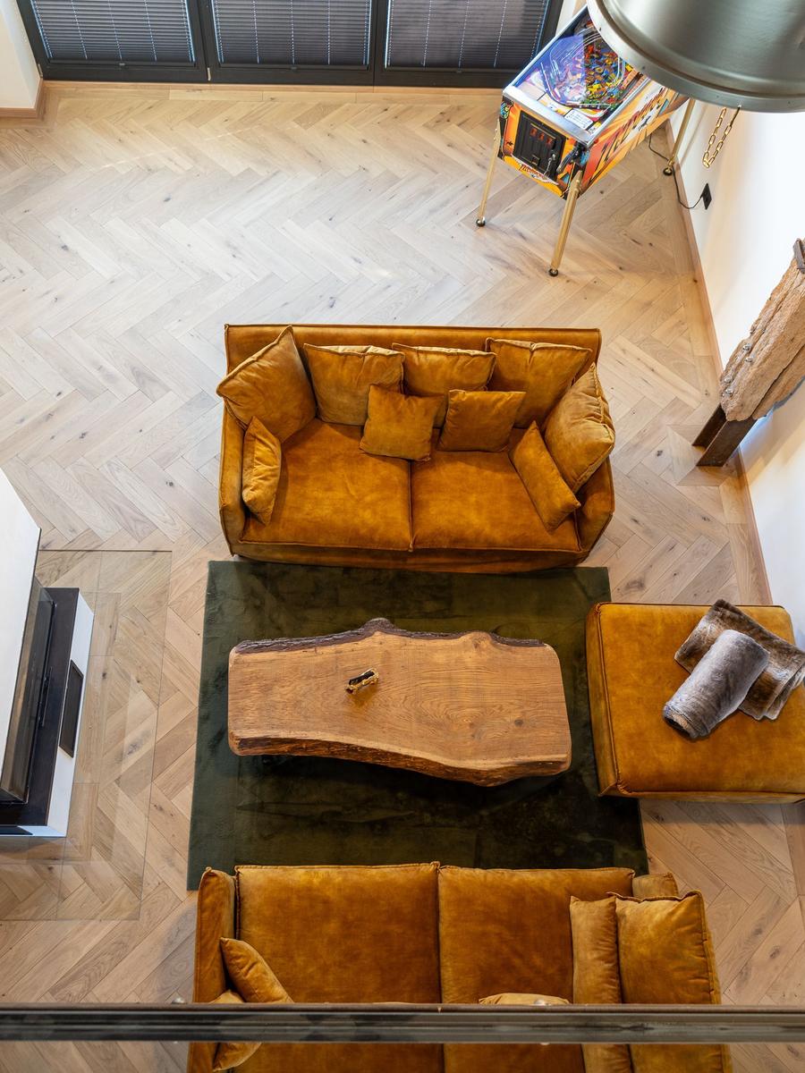 Zwei goldfarbene Sofas, ein Holztisch und ein Pinballautomat auf hellem Parkettboden.