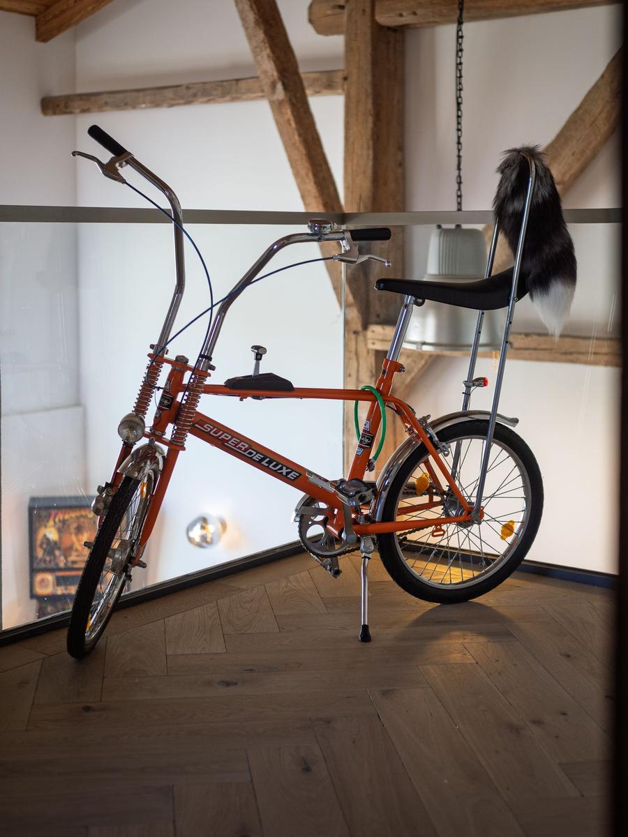 Ein orangefarbener Super Deluxe Fahrrad steht im Raum mit Holzboden und Balken.