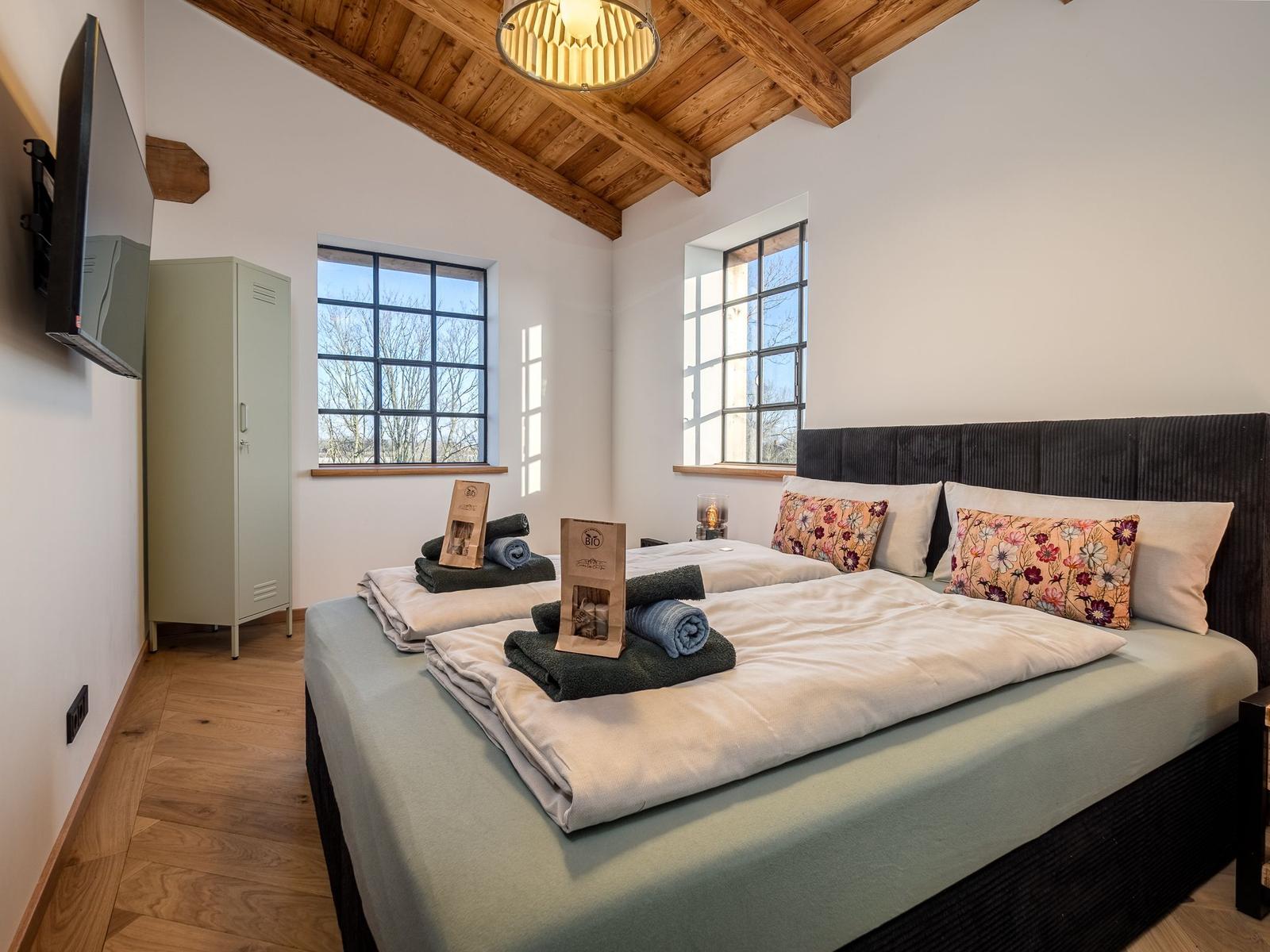 Schlafzimmer mit Doppelbett, Holzboden und zwei Fenstern. Wandmontierte TV und Kleiderschrank.