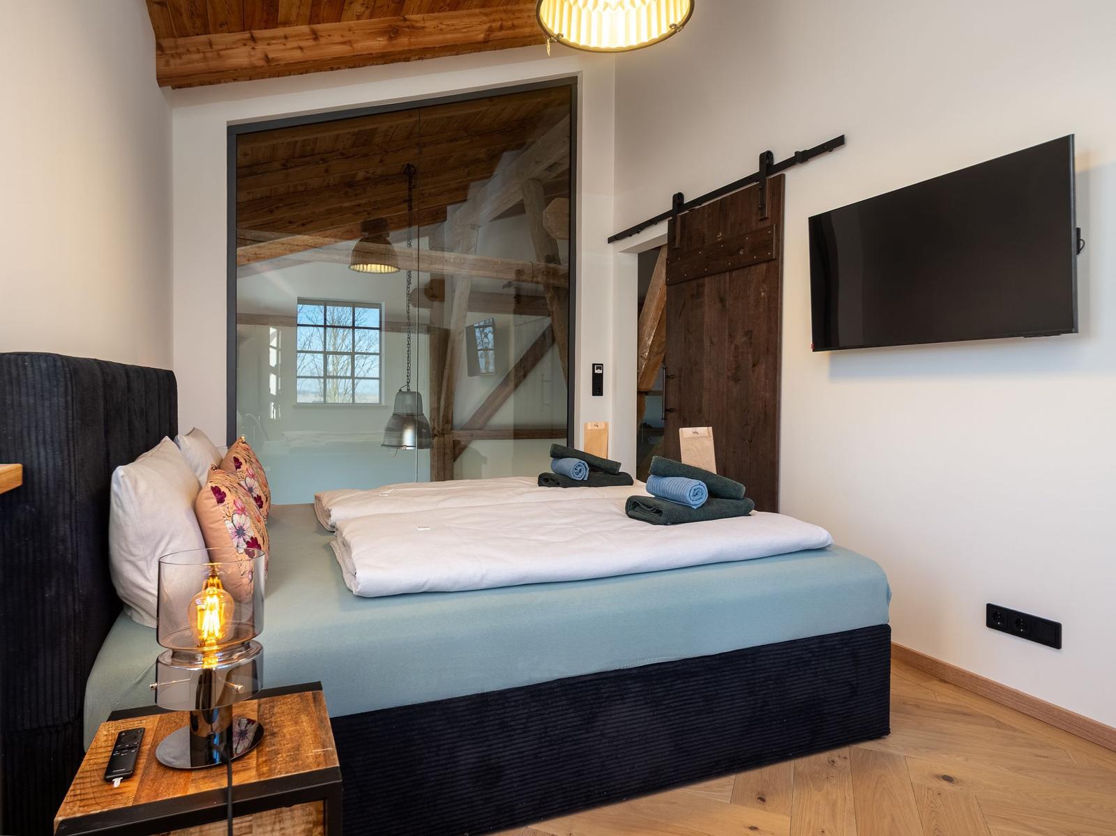 Schlafzimmer mit Bett, Fernseher und Holzboden. Große Glaswand und hölzerne Tür.