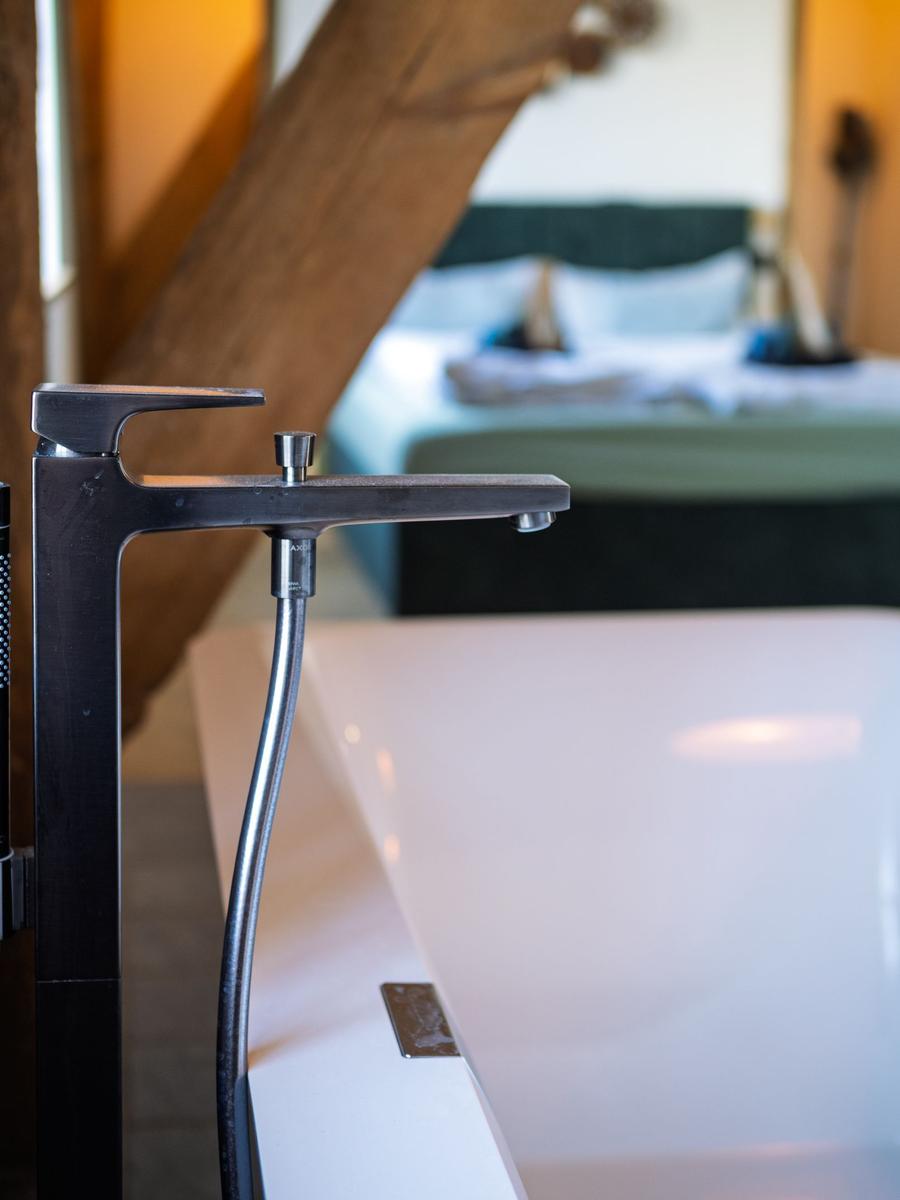 Duschbrause über weißem Badewannenrand, im Hintergrund Bett mit dunklem Kopfteil.