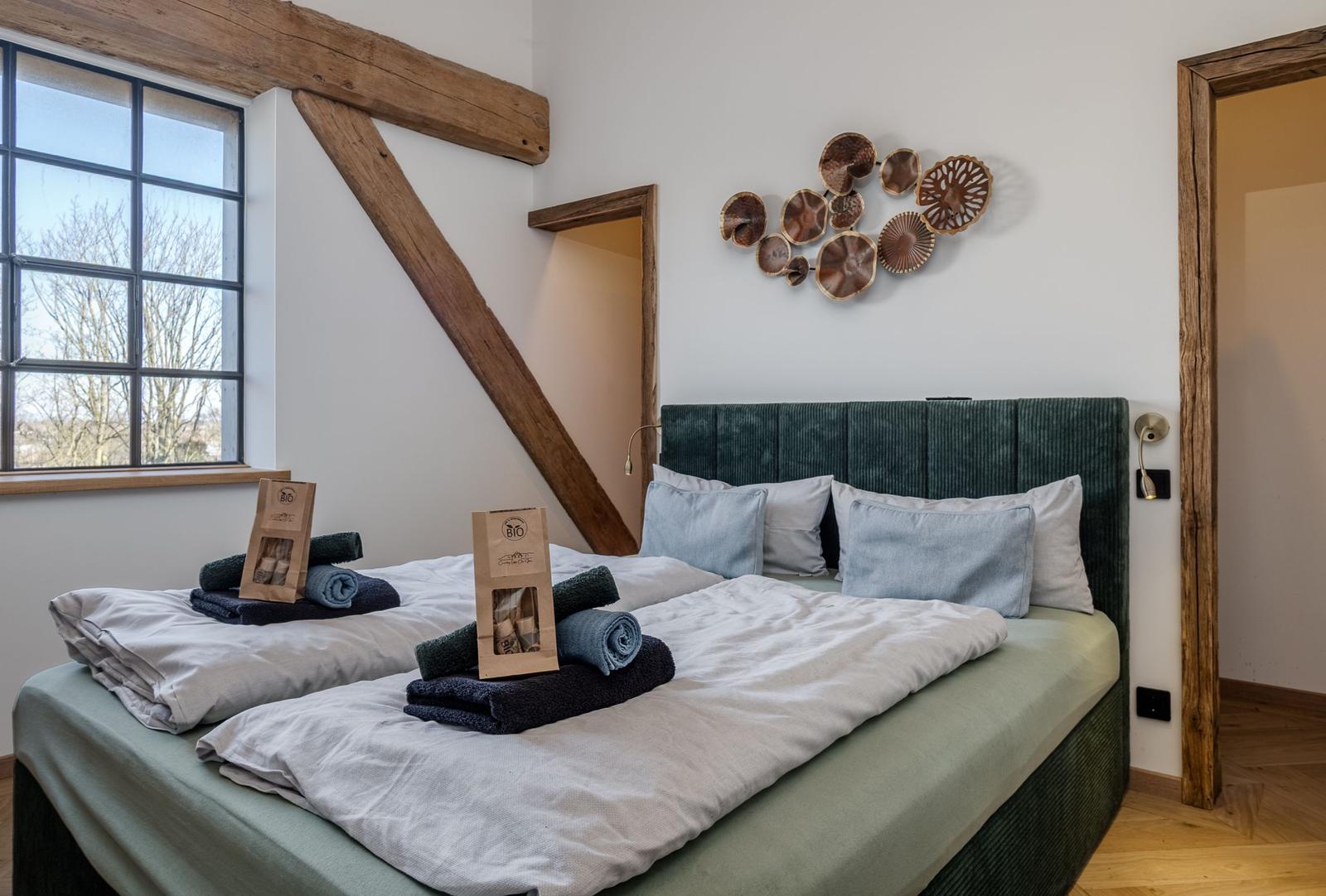 Doppelbett mit grünem Kopfteil, Holzbalken und Fenster mit Blick.