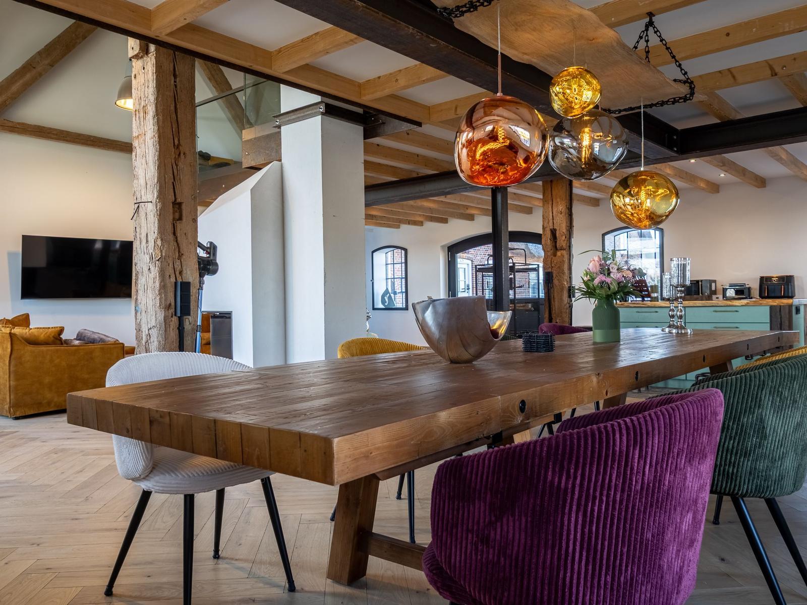 Essbereich mit massivem Holztisch und bunten Stühlen unter Holzbalkendecke.