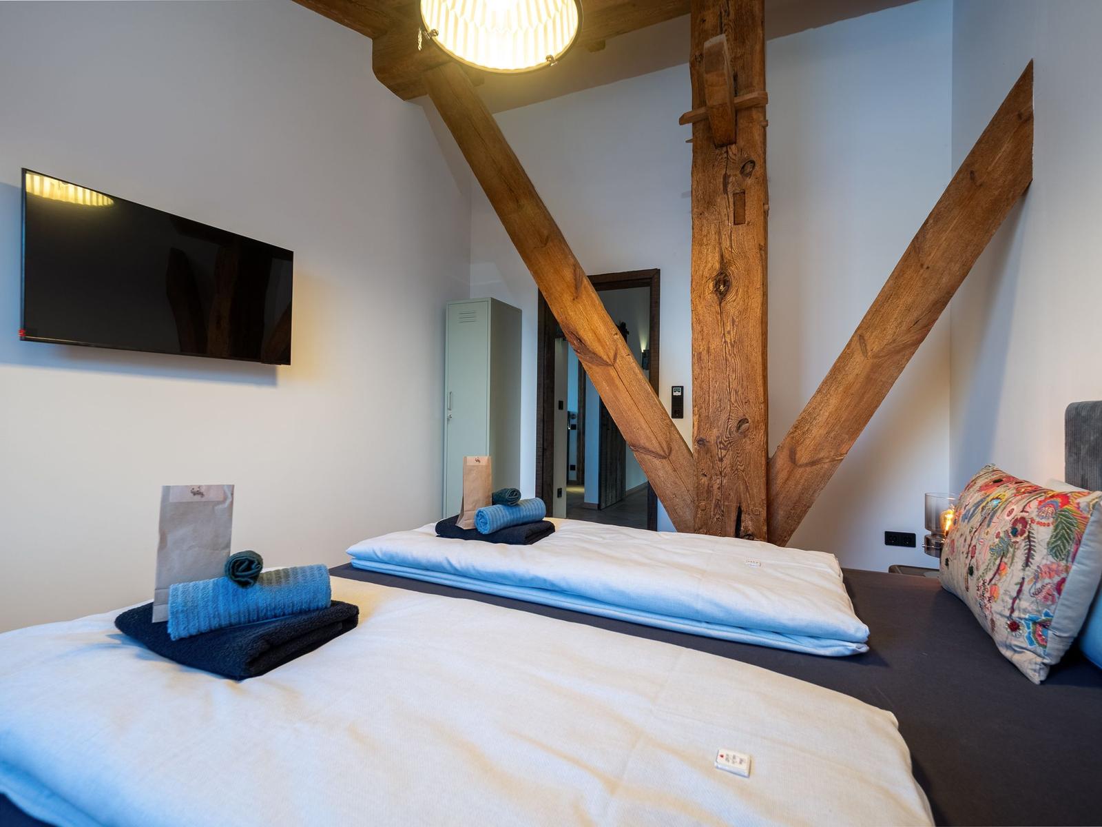 Schlafzimmer mit Holzbalken, Bett, Fernseher und Handtüchern.