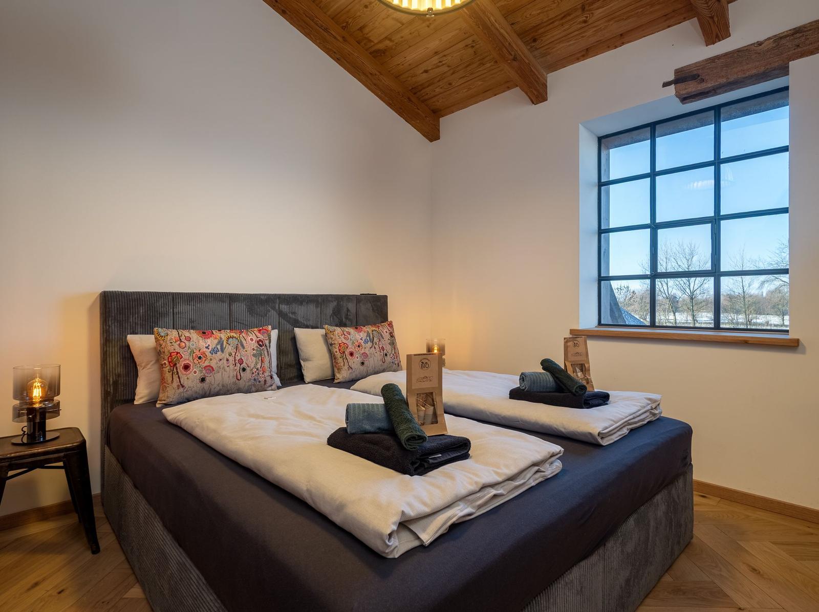 Schlafzimmer mit Doppelbett, Holzbalkendecke und großem Fenster.