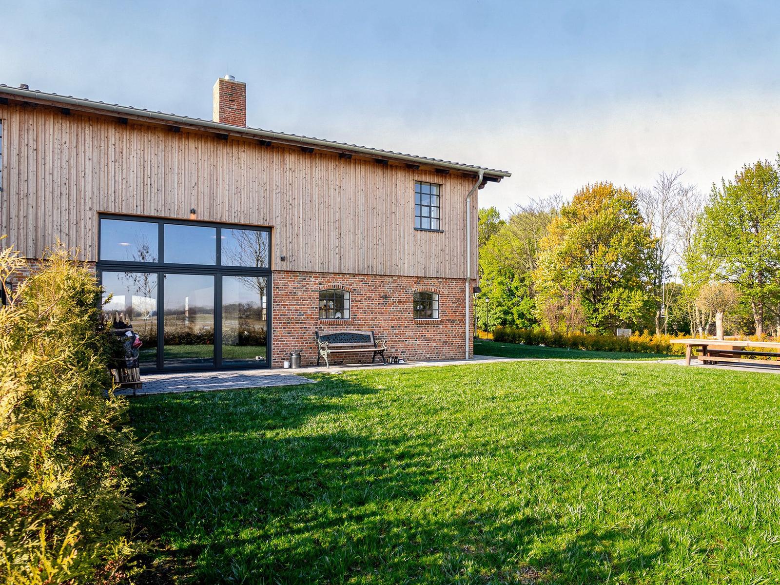 Haus mit Holzfassade und Backsteinbasis, großes Fenster, Garten und Bänke.