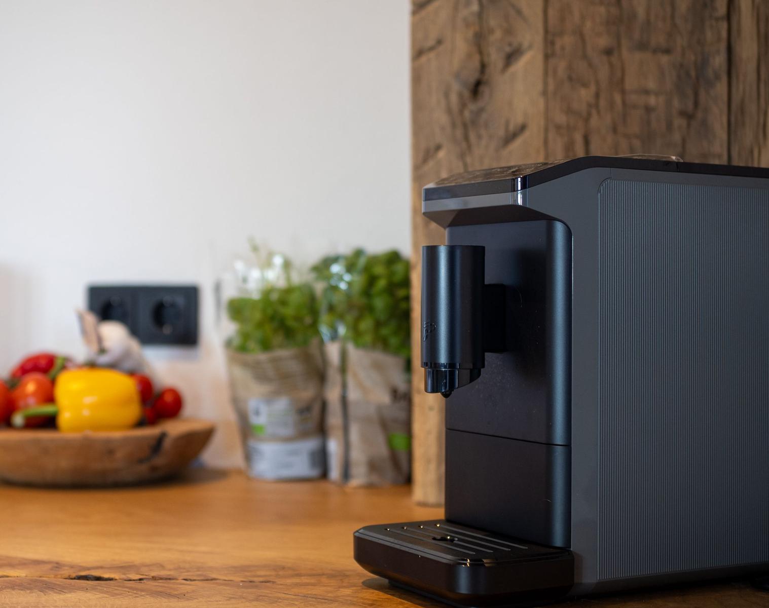 Eine schwarze Kaffeemaschine steht auf einer Holzoberfläche. Im Hintergrund sind Gemüse und Pflanzen zu sehen.