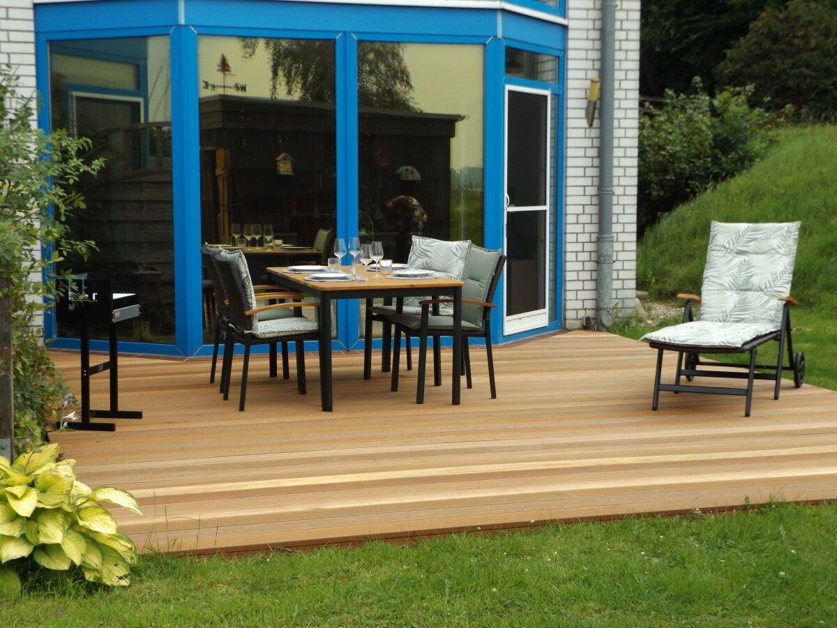 Deck with table, chairs, and lounge chair in front of blue house with glass facade.