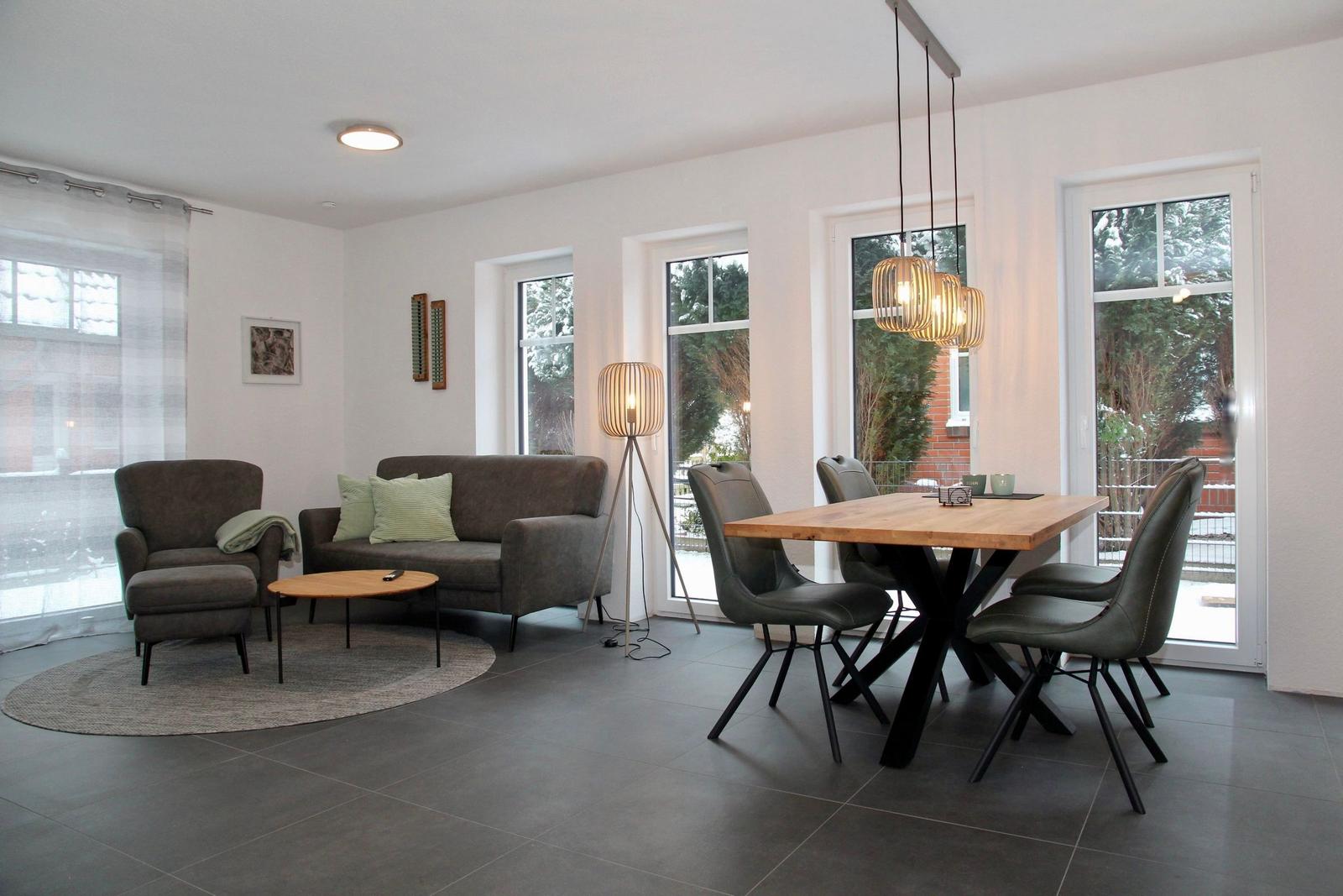 Wohn- und Essbereich mit Sofa, Sessel, Tisch und Stühlen. Fenster und Tür zu Garten.