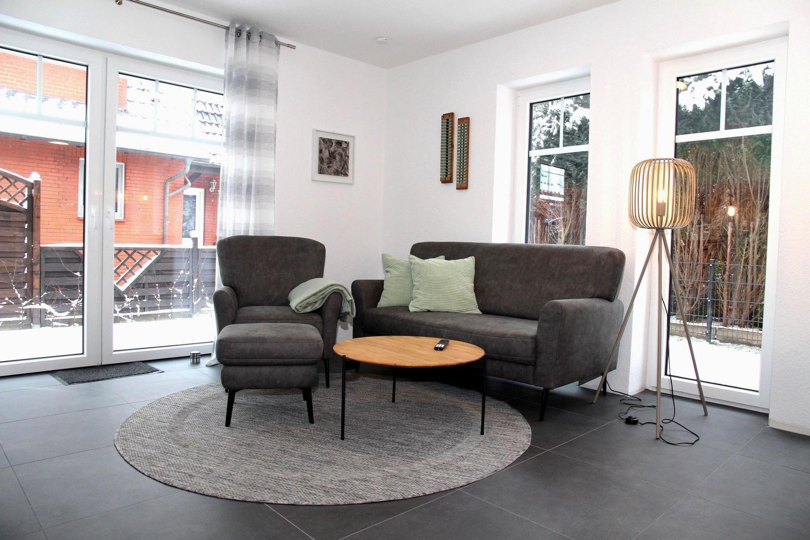 Wohnzimmer mit Sofa, Sessel und runden Teppich. Große Fenster mit Blick nach draußen.
