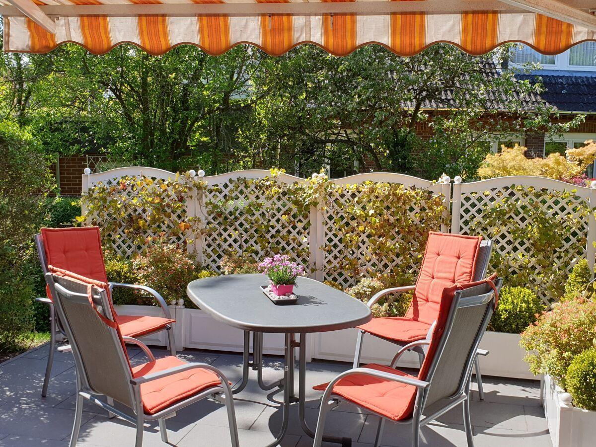 Terrasse mit Tisch, vier Stühlen und Sonnenschutz. Hintergrund: weißer Zaun und Pflanzen.