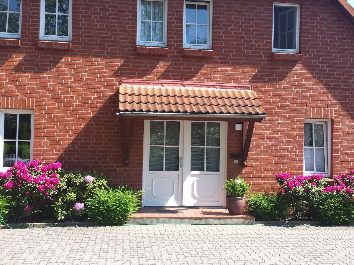 Rotes Backsteinhaus mit weißer Eingangstür und blühenden Pflanzen vor dem Eingang.
