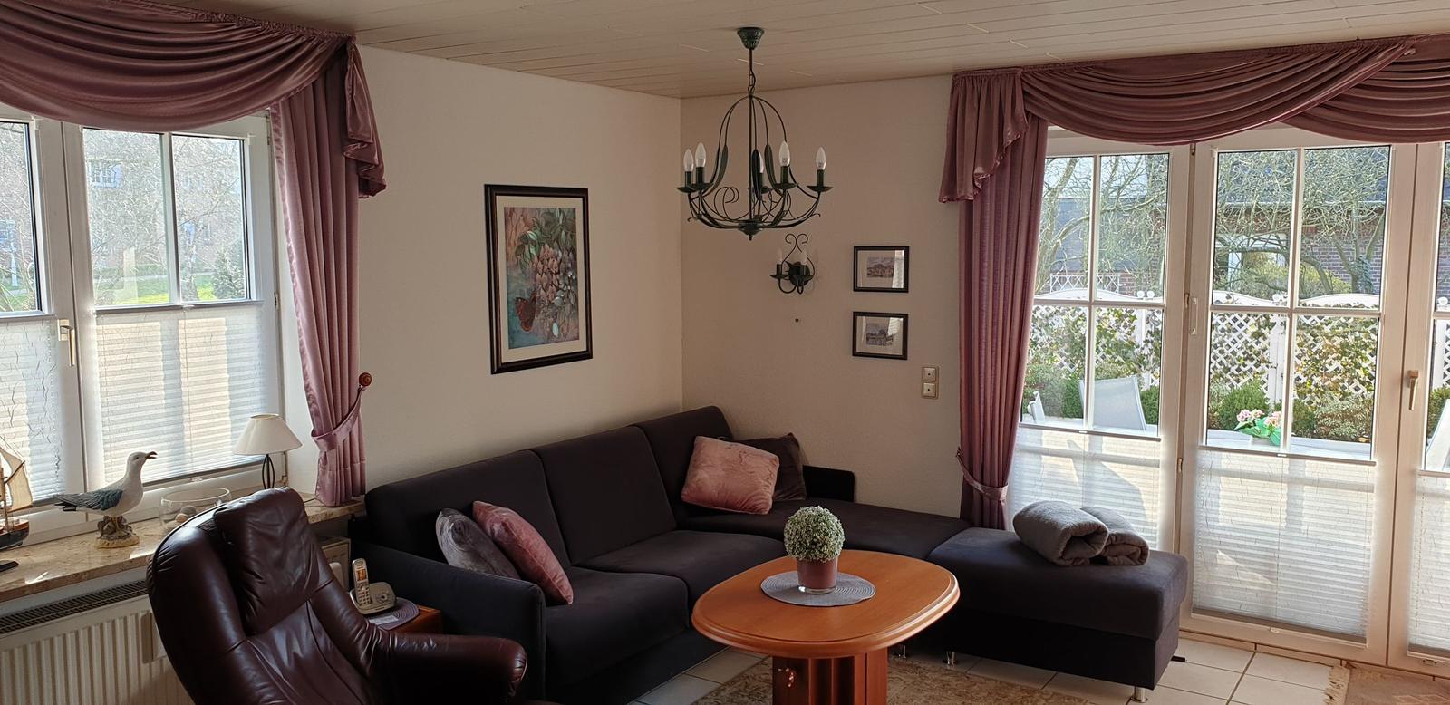 Wohnzimmer mit Sofa, Sessel und Tisch. Fenster mit Vorhängen und Blick auf Terrasse.