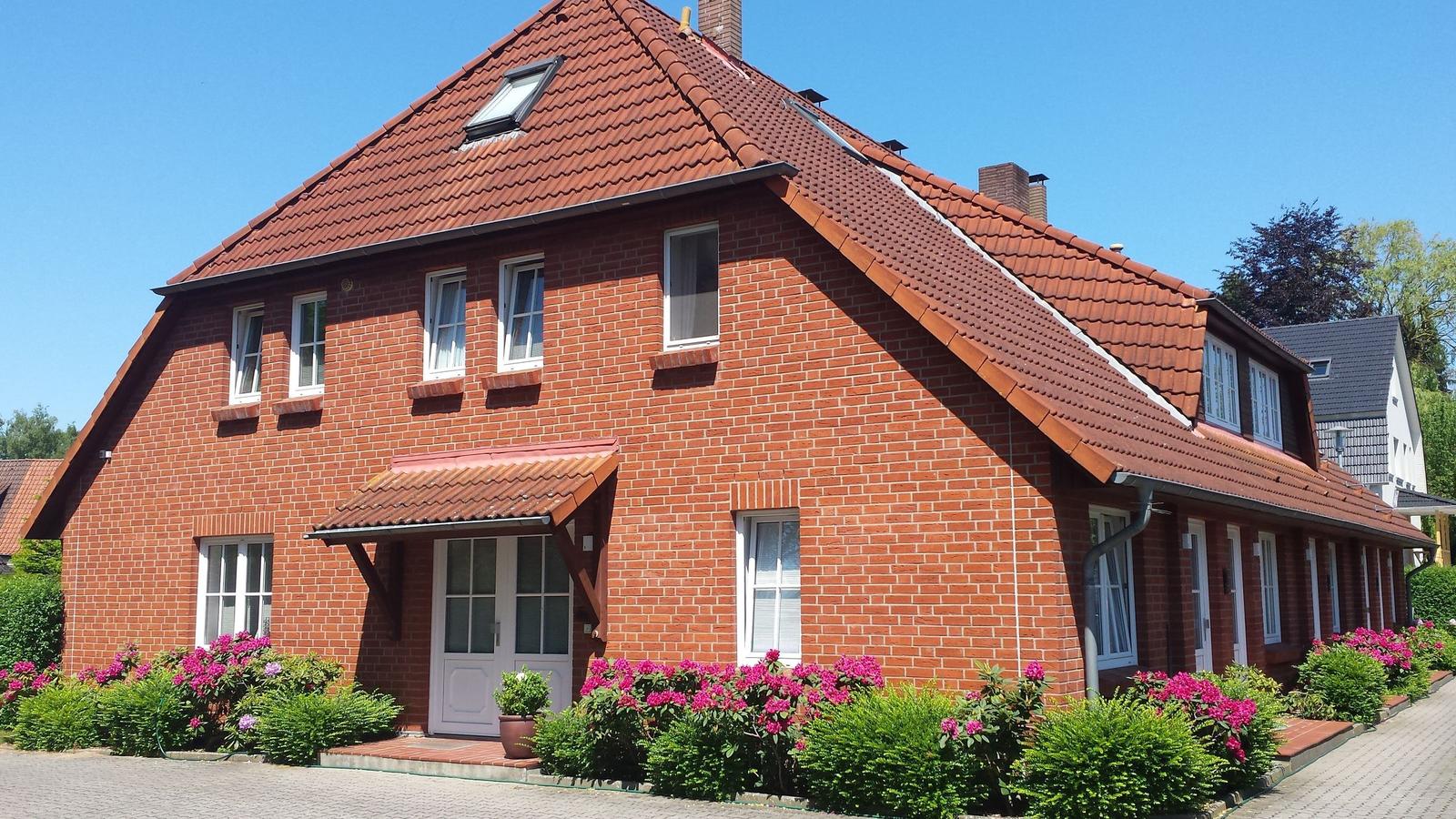 Rotes Backsteingebäude mit Dachgauben und blühenden Sträuchern vor der Tür.
