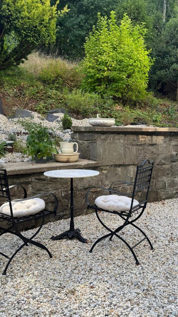 Garden with table and two chairs on gravel, stone wall and plants in background.