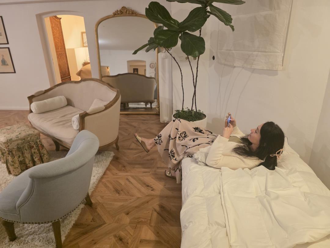 A person lies on a bed looking at their phone. In the background, there is a sofa and a mirror.