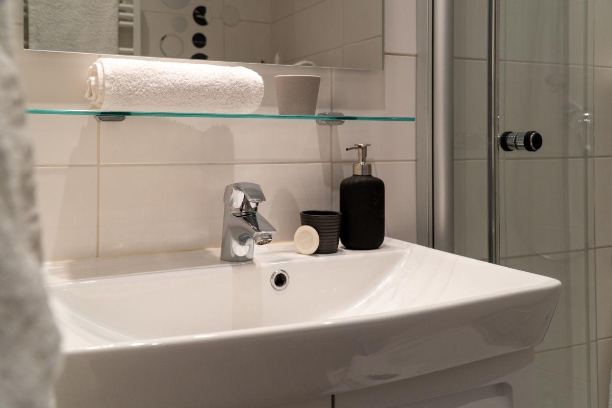 Bad mit Waschbecken, Spiegel und Dusche. Auf der Ablage liegt ein Handtuch.