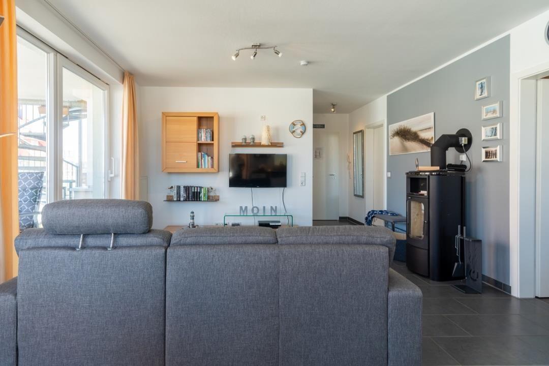 Wohnzimmer mit Sofa, Fernseher und Kaminofen. Große Fenster mit Blick auf Balkon.