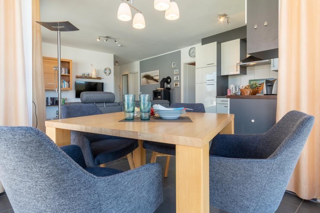 Essbereich mit Holztisch und blauen Stühlen. Küche und Wohnzimmer im Hintergrund.