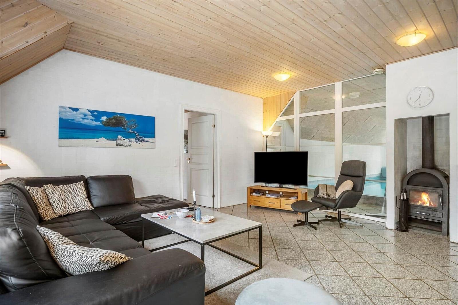 Wohnzimmer mit Sofa, Fernseher und Kaminofen. Glaswand zeigt Pool.