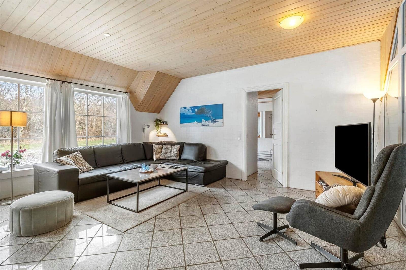 Wohnzimmer mit Sofa, Sessel und Fernseher. Holzdecke und große Fenster.