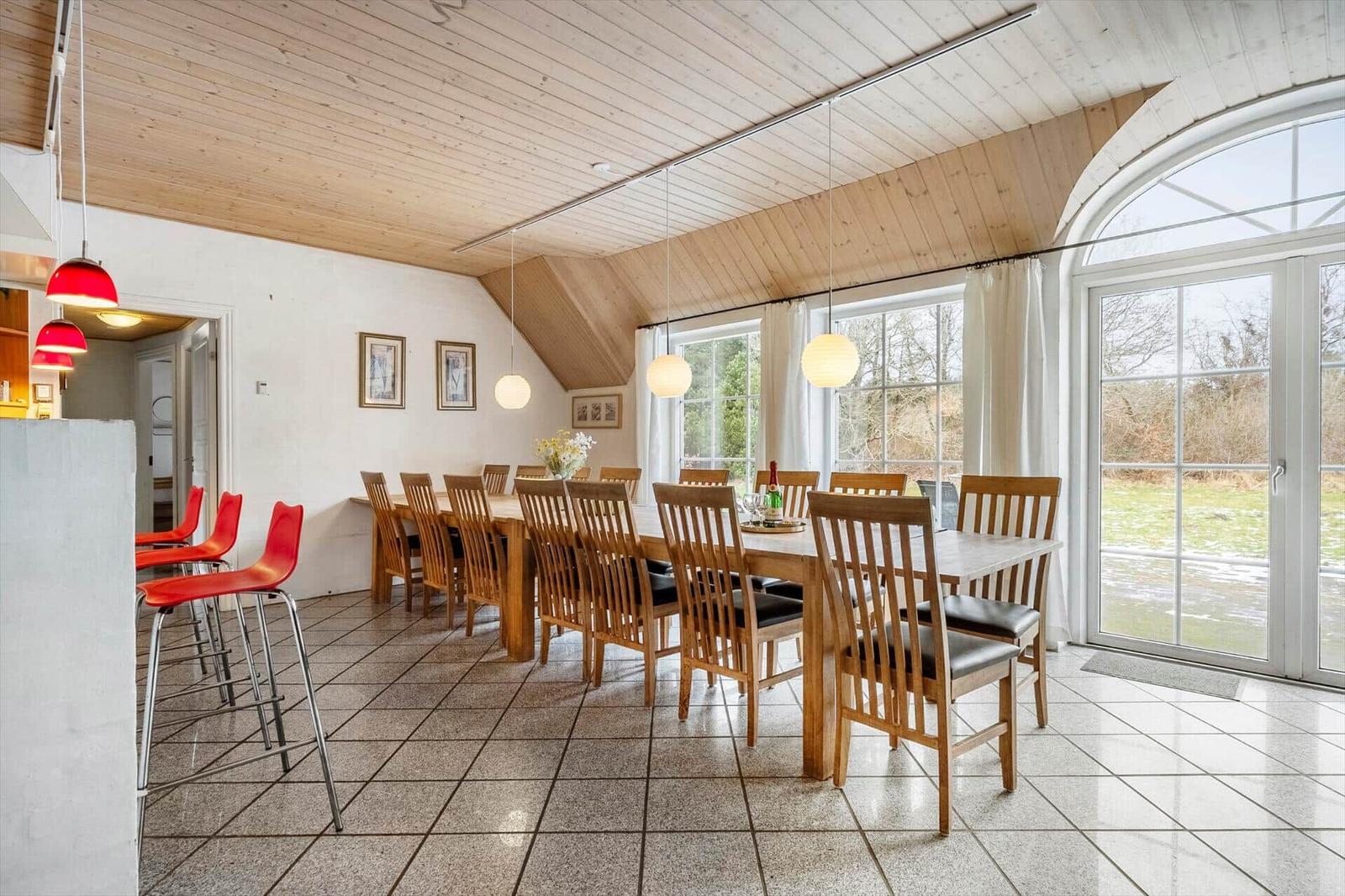 Esszimmer mit Holztisch, Stühlen und großem Fenster. Rote Barhocker und helle Decke.