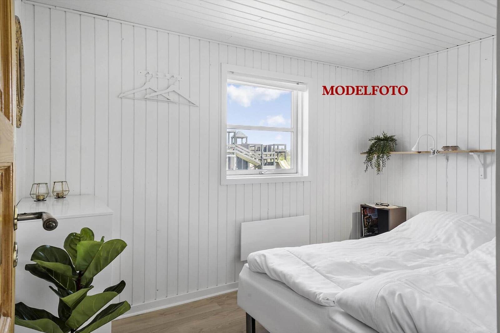 Schlafzimmer mit weißem Bett, Fenster und Holzboden. An der Wand hängt ein Kleiderhaken.