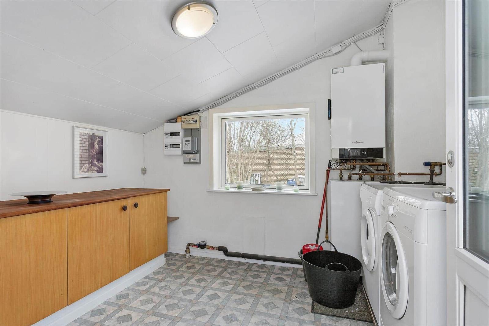 Room with washing machine, boiler, and wooden cabinet. Window overlooking outdoor area.