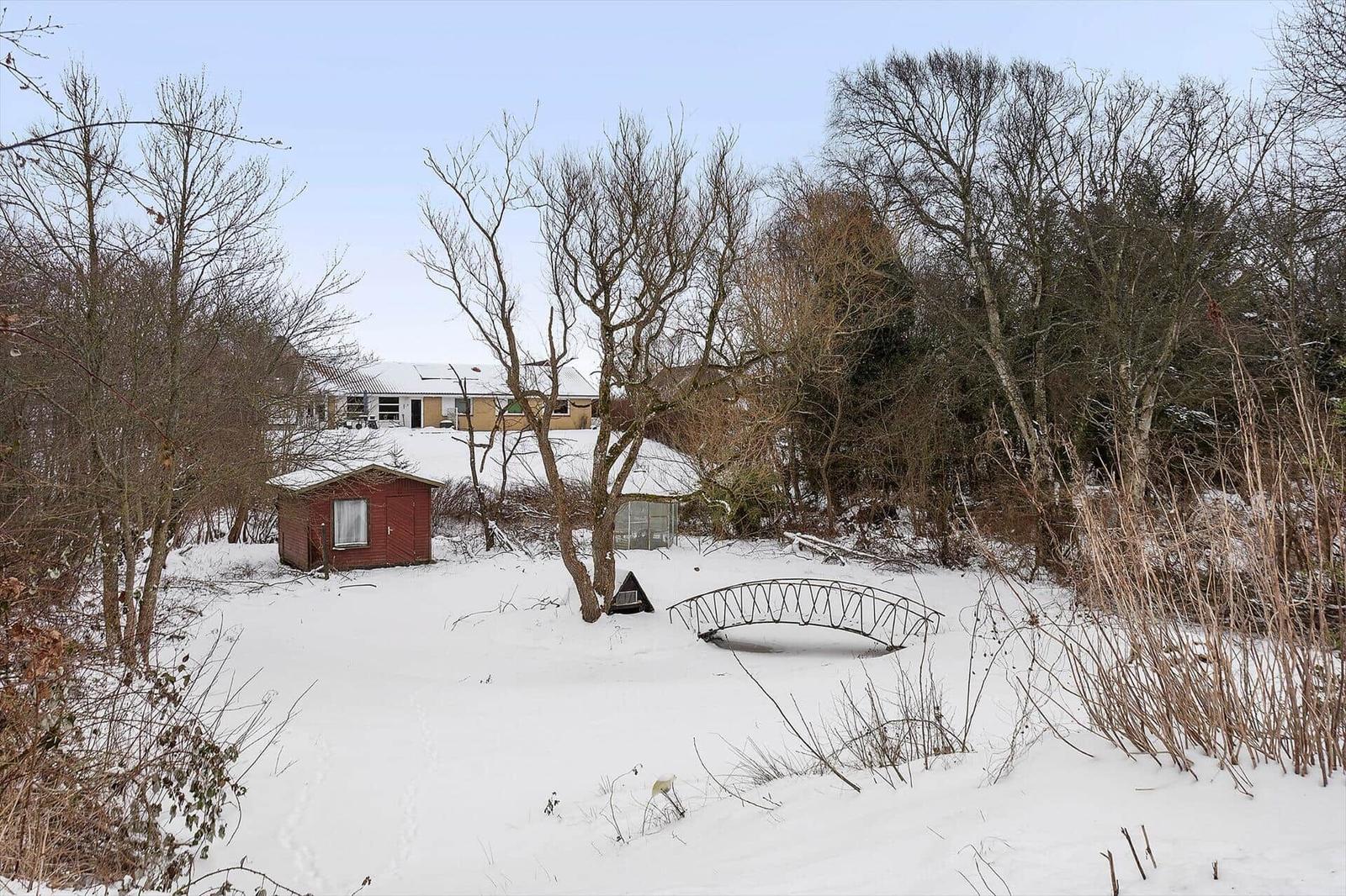 Snowy landscape with house, red shed, and metal bridge among trees.