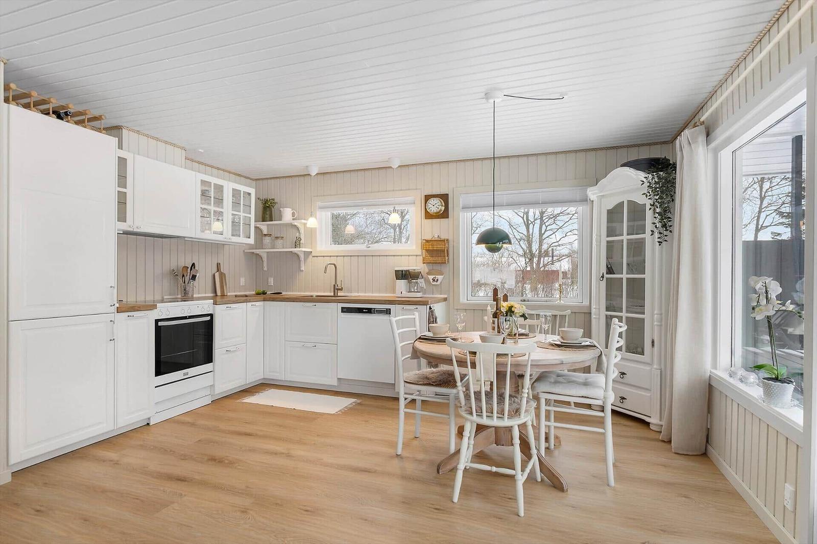 Kitchen with dining table, white cabinets, and wooden floor. Large windows and light walls.