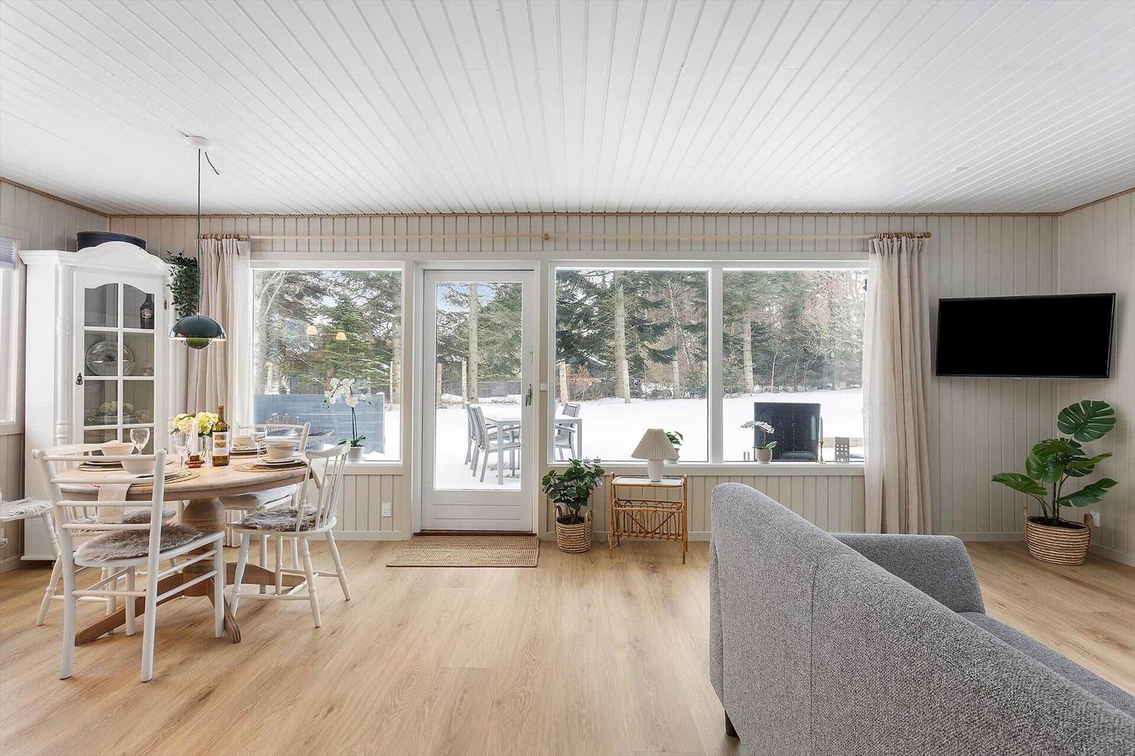 Living and dining area with wooden floor, large windows and TV. Outside snow and trees.