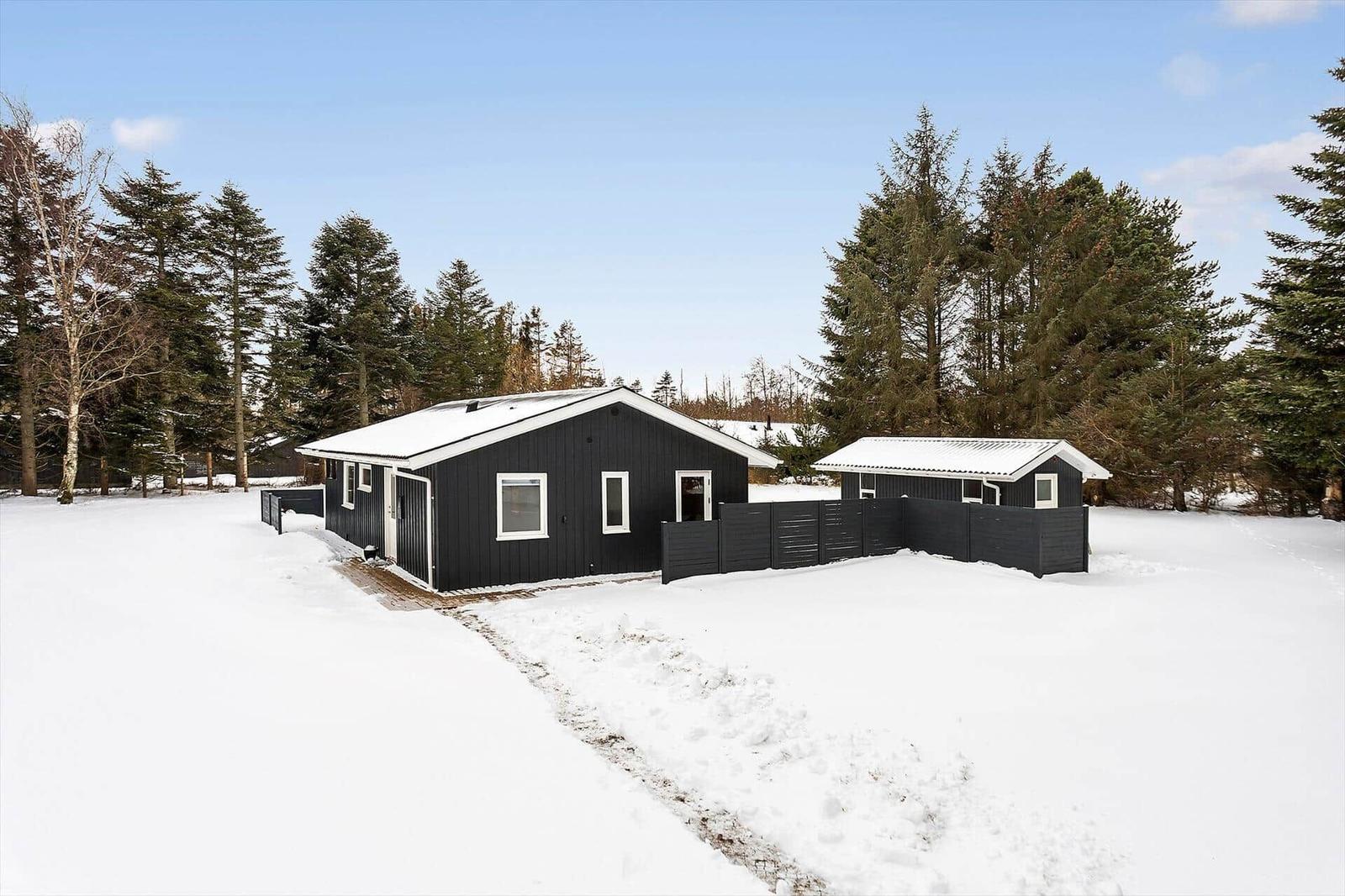 Black wooden house with snow-covered roof and surrounding trees.