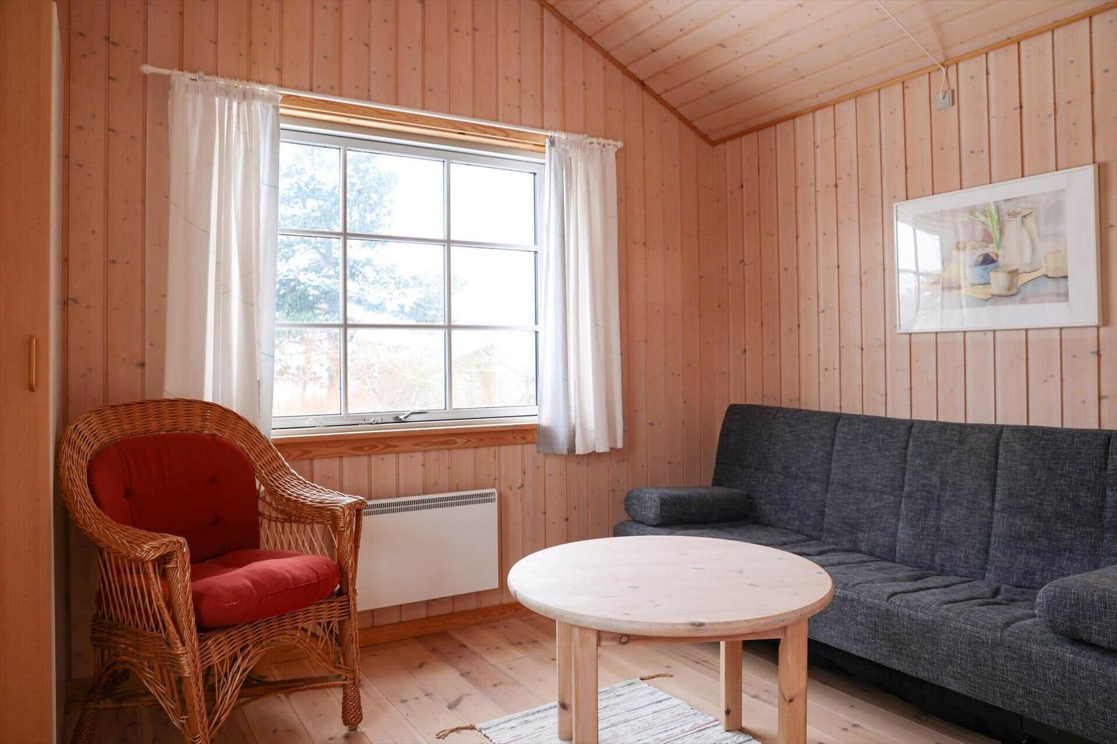 Wohnzimmer mit Sofa, Rattan-Stuhl und runden Tisch. Holzwände und Fenster mit weißen Vorhängen.