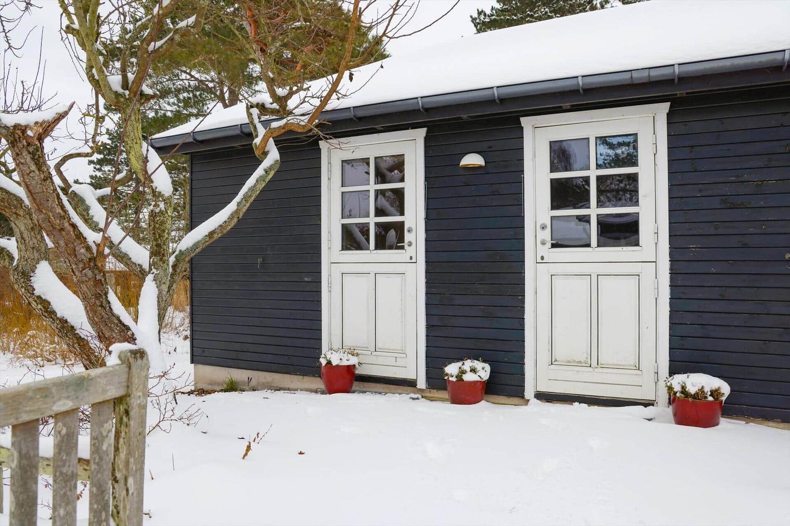 Schwarzes Holzhaus mit zwei weißen Türen und Schneebedeckten Pflanzen.
