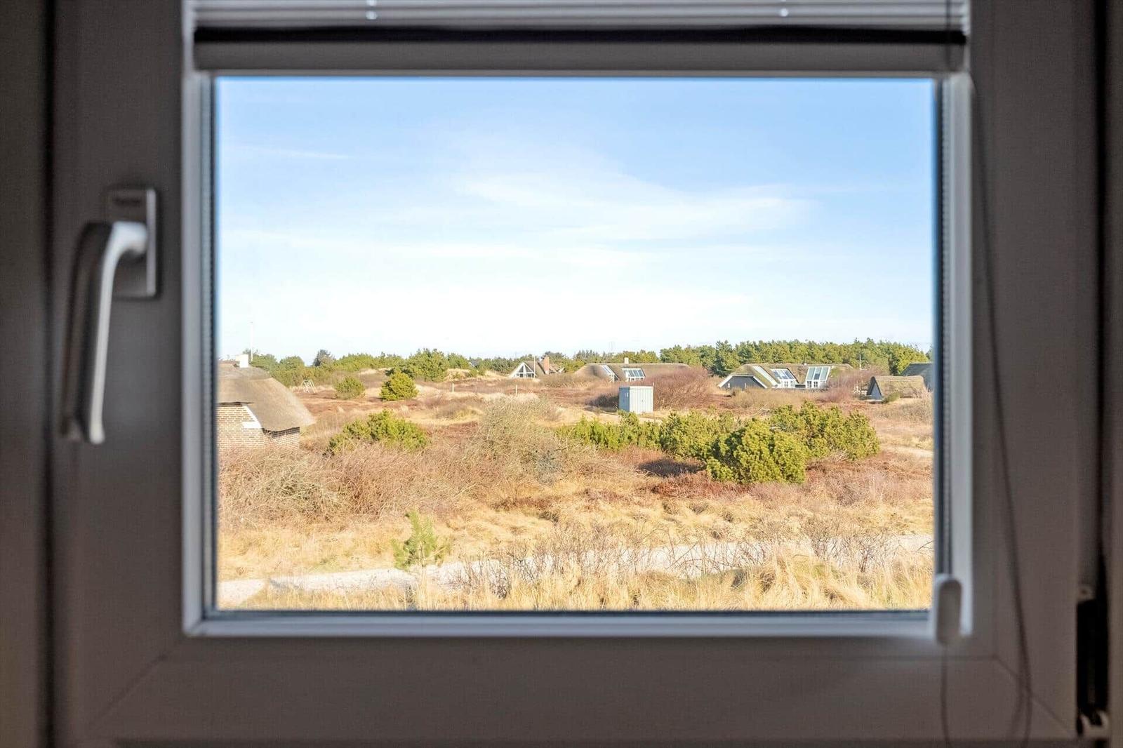 Durch das Fenster blickt man auf eine trockene Heidelandschaft mit Häusern und Bäumen.