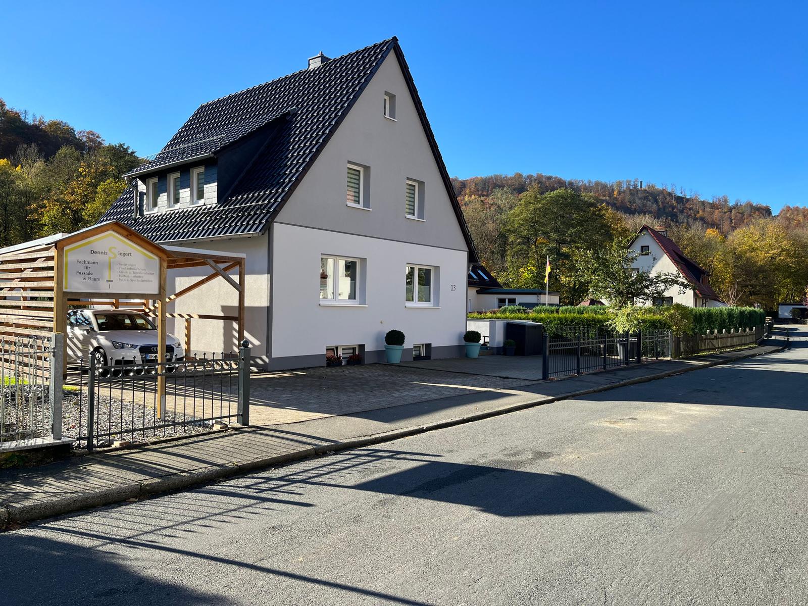 Haus mit dunklem Dach und weißer Fassade, gepflastertes Vorfeld und begrünte Umgebung.