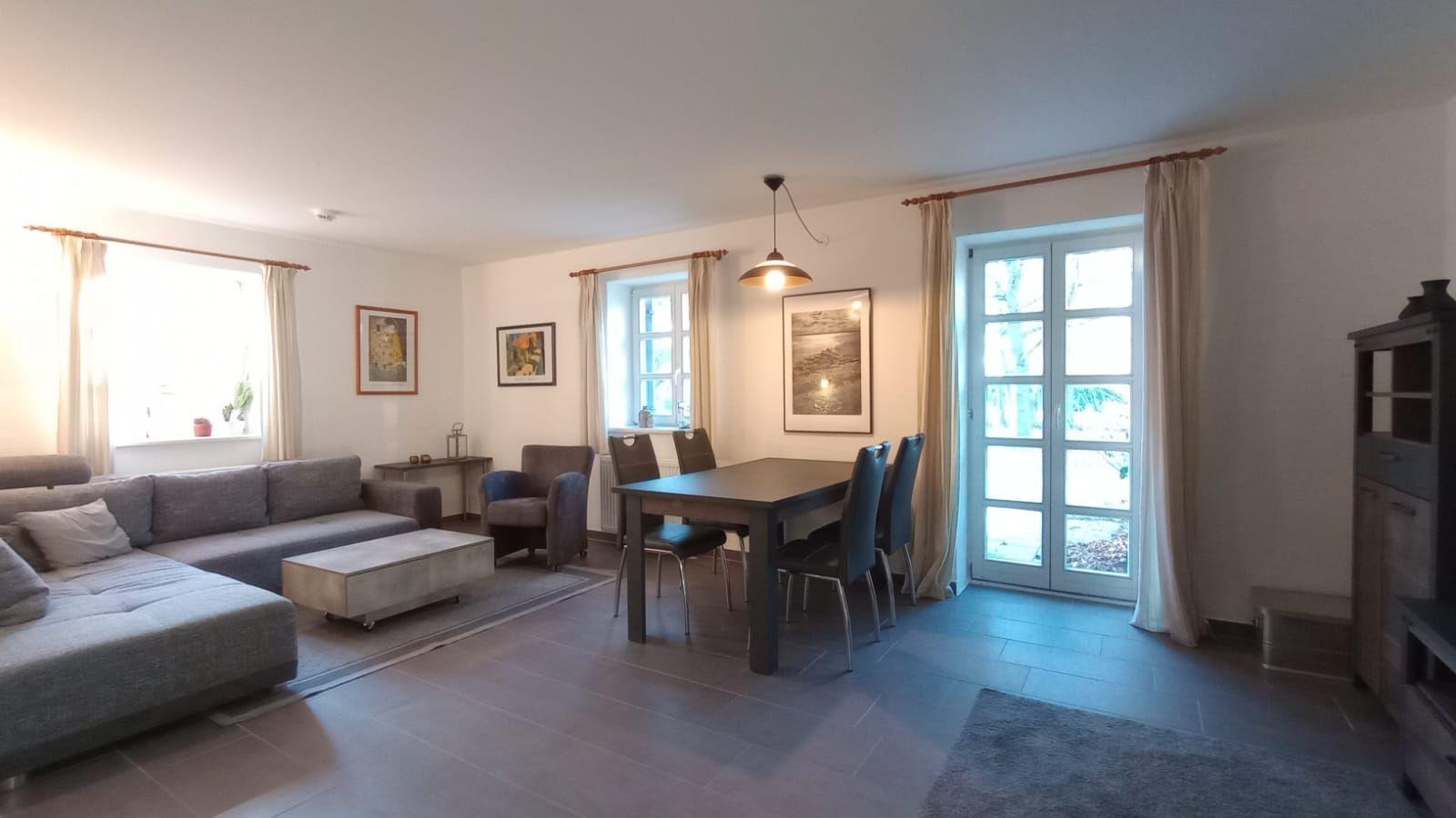 Wohn- und Essbereich mit Sofa, Tisch und Stühlen. Fenster und Glastür mit Vorhängen.