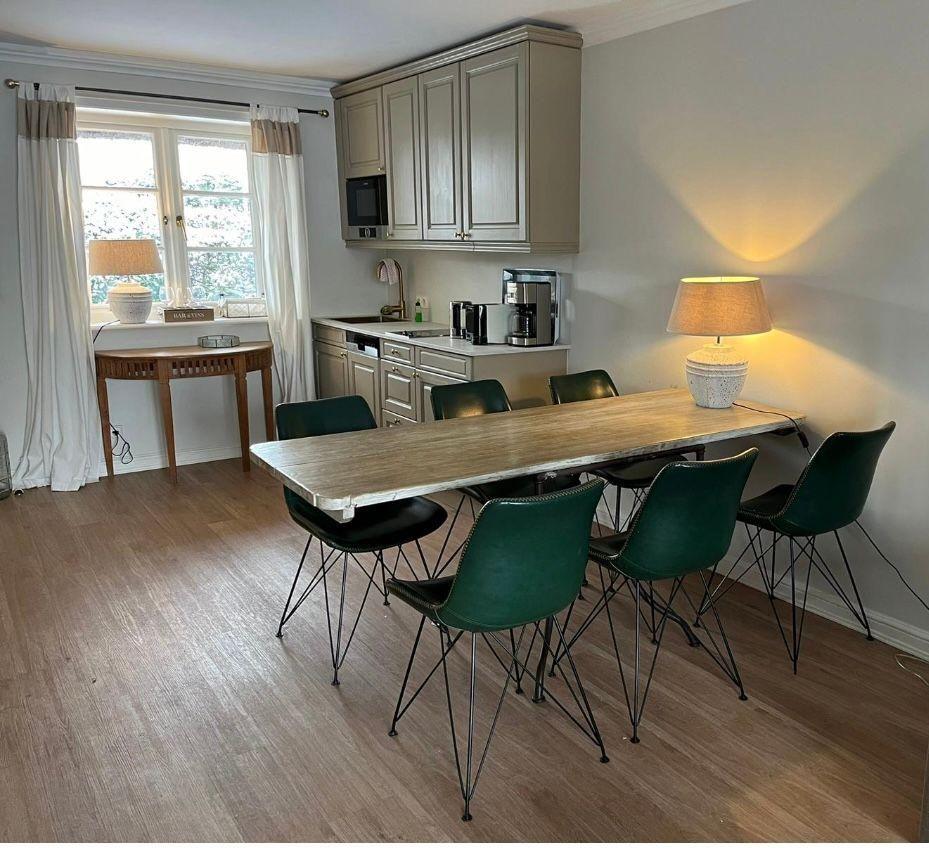 Essbereich mit langer Tisch und grünen Stühlen, Küchenzeile und Fenster mit Vorhängen.