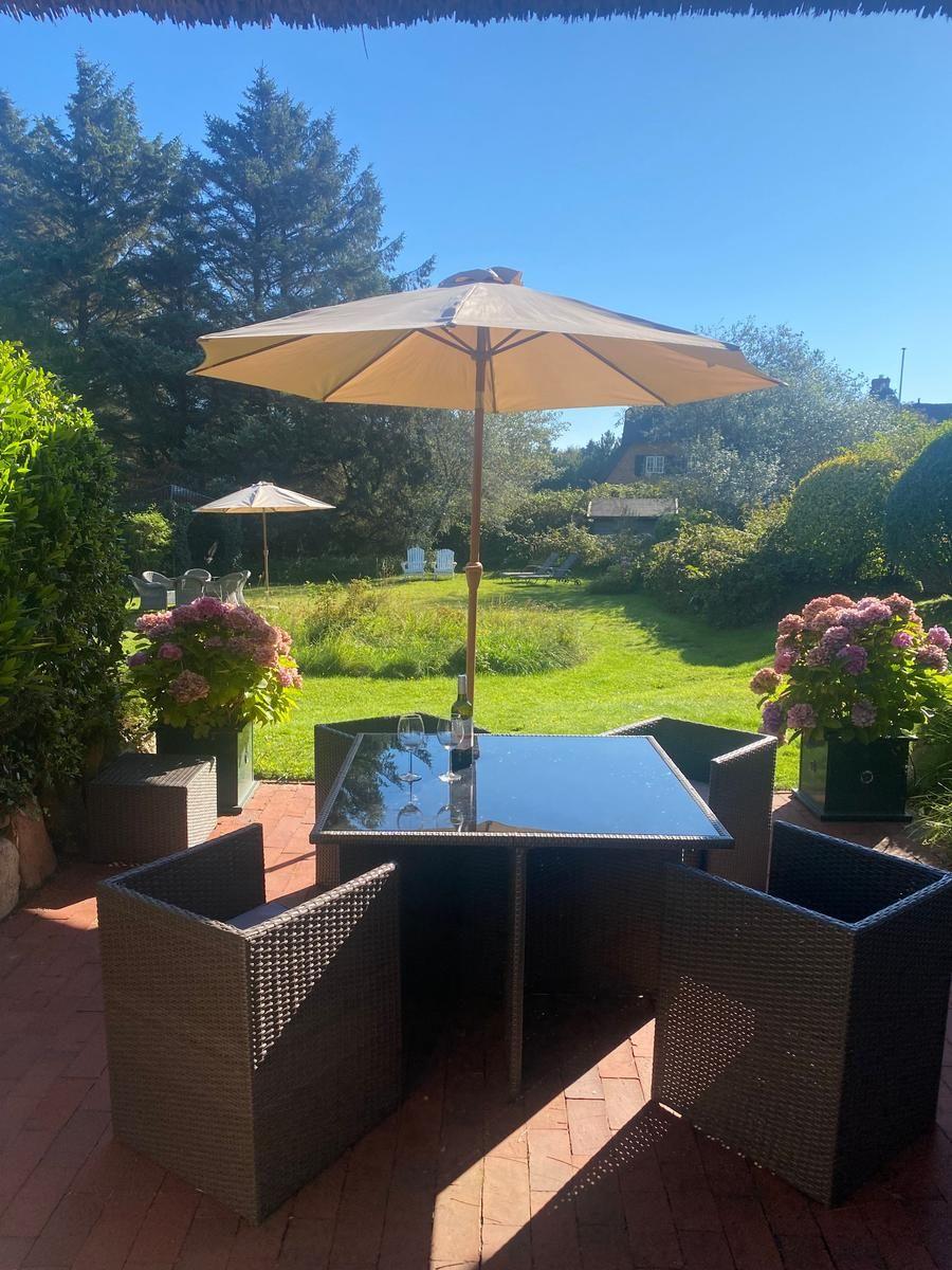 Terrasse mit Tisch, Stühlen und Sonnenschirm. Grünflächen und Blumen im Hintergrund.