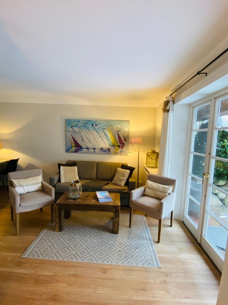 Wohnzimmer mit Sofa, zwei Sesseln und Glas-Tür zu Außenbereich.